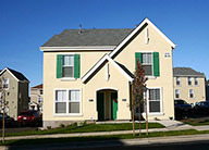 Two-story beige stucco townhome-style unit with gabled roof and green doors and shutters.
